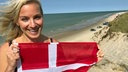 eine Frau mit langen blonden, im Wind wehenden Haaren hält fröhlich lachend eine rote Stoffflagge mit weißem Kreuz vor ihrem Oberkörper, dahinter Dünen, ein Strand, auf dem Autos parken, und die grün-türkise Nordsee