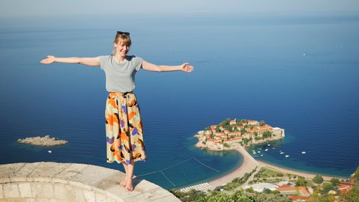 junge blonde Frau in hellgrauem T-shirt und orange-lila gemustertem wadenlangen Rock steht barfuß und mit ausgebreiteten Armen auf einer hellen Steinmauer, dahinter weiter unten blaues Meer mit Küste und einer kleinen Insel