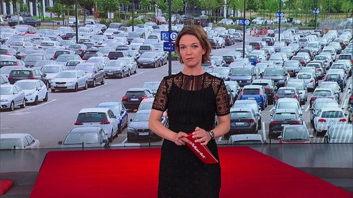 Anna Planken steht im Markt-Fernsehstudio. Im Hintergrund ist ein Parkplatz abgebildet. 