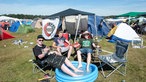 Drei Junge Herren auf einem Campingplatz, die Füße in ein Kinderschwimmbecken getaucht, die Sonne scheint, die Stimmung wirkt ausgelassen