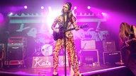 Kate Nash spielt nun wieder Gitarre und ist in voller Größe und in lilanem Licht auf der Bühne mit ihrer Band zu sehen.