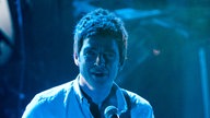 Noel Gallagher ist in blaues Licht getaucht und sieht genau in die Kamera.