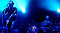 Gitarrist spielt Gitarre und singt in ein Mikrofon, schräg links von Ihm spielt ein Mann das Schlagzeug, im Hintergrund sieht man blau strahlende Scheinwerfer