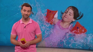 Daniel Aßmann steht im Servicezeit-Fernsehstudio. Hinter ihm sieht man ein kleines Mädchen im Schwimmbad.