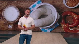 Daniel Aßman steht im Servicezeit-Studio. Im Hintergrund sieht man eine bayrische Weißwurst.