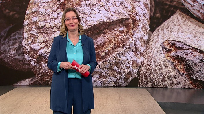 Yvonne Willicks steht im Servicezeit-Studio. Hinter ihr sind mehrere Laibe Brot abgebildet.