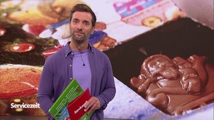 Der Moderator steht im Studio. Im Hintergrund ist ein Adventskalender zu sehen.