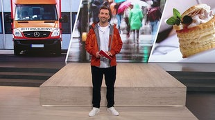 Moderator Daniel Aßmann steht im Servicezeit-Studio, hinter ihm sind Bilder von einem Rettungswagen, Regenjacken und Gebäck eingeblendet.