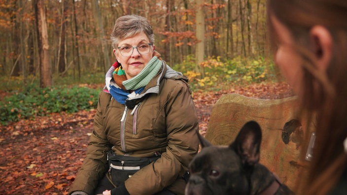 Eine Frau mit kurzen Haaren und einer braunen Jacke sitzt mit einem schwarzen Hund im Wald auf einer Bank und unterhält sich mit einer anderen Frau