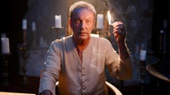 Udo Kier sitzt am dunklen Spielfimset an einem Tisch, hält ein großes brennendes Streichholz hoch und schaut mit seinen markanten stahlblauen Augen in die Kamera