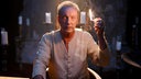 Udo Kier sitzt am dunklen Spielfimset an einem Tisch, hält ein großes brennendes Streichholz hoch und schaut mit seinen markanten stahlblauen Augen in die Kamera