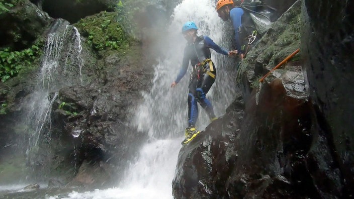 Tamina Kallert im Neoprenanzug und mit Helm steht auf einem Felsen inmitten eines Wasserfalls