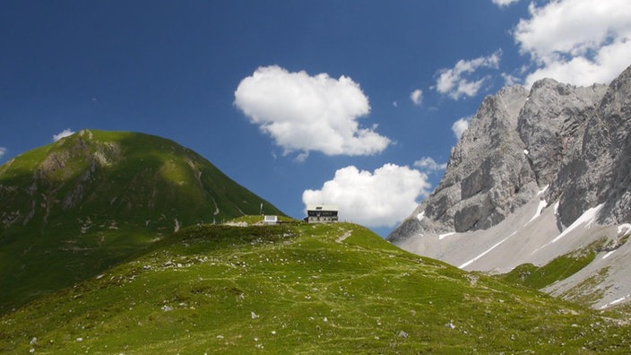 Die Kemptner Hütte auf einer grünen Bergkuppe, daneben steiles Bergmassiv