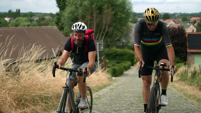 Daniel Aßmann (l) und Radfahrprofi Johan Meuseeuw fahren auf Rennrädern eine Straße bergauf