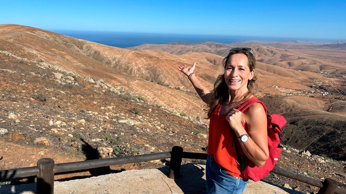 Tamina Kallert steht auf einem Aussichtspunkt und zeigt über kahle Hügel auf das entfernte Meer