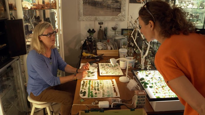 Anne Wilmes im Gespräch mit Maren Lohmeyer (l) an einem Tisch mit ausgebreiteten Meerglasstücken