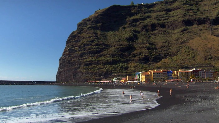 Schwarzer Sandstrand, endet mit einem kleinen Ort vor dunkler, hoher Felswand