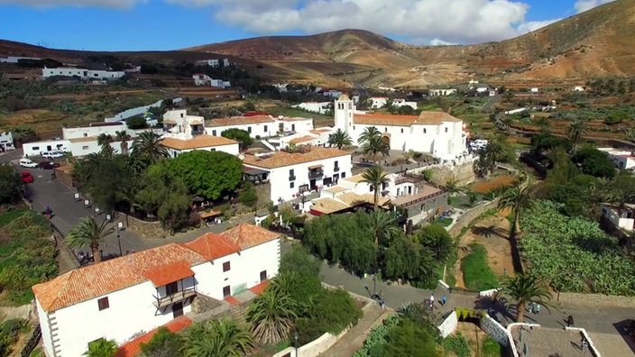 Ort mit weißen Häusern und Kirche, im Hintergrund bergige Landschaft