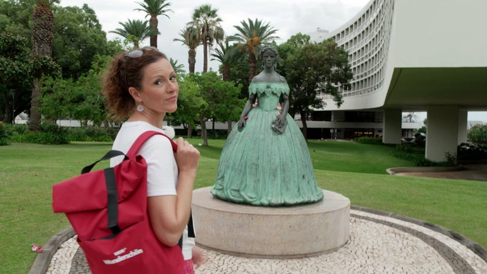 Anne Willmes steht vor einer Statue der Kaiserin Elisabeth I. in einem Park mit Palmen