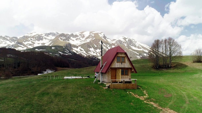 Kleines Holzhaus mit Giebeldach auf einer Wiese, im Hintergrund teils schneebedeckte, hohe Berge