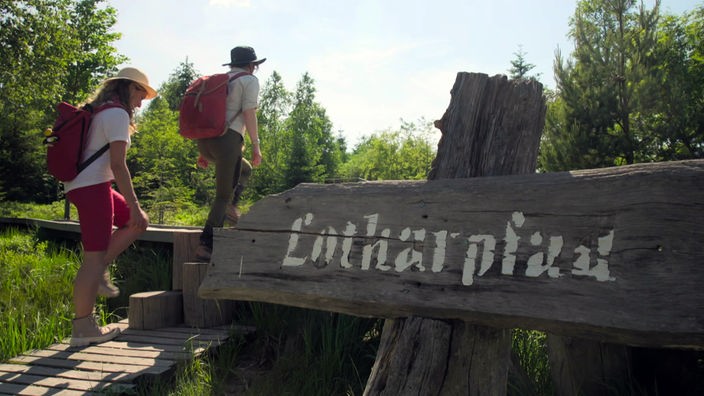 Zwei Wanderer auf dem Lotharpfad, der im Vordergrund mit einem Holzschild angekündigt wird