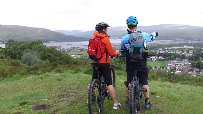 Tamina Kallert und eine weitere Person mit Mountainbikes stehen auf einem Hügel und schauen auf die darunter liegende Landschaft 