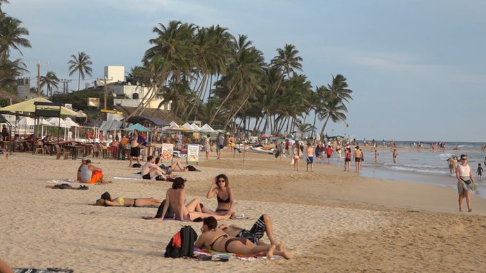 Badegäste an einem Sandstrand mit Gastronomie und Palmen