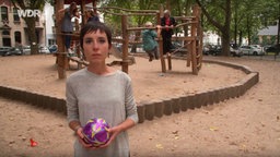 Samay Böll steht auf einem Spielplatz in der Kölner Südstadt und hält einen lila-farbenen Ball in der Hand, Böll folgen Videowalk