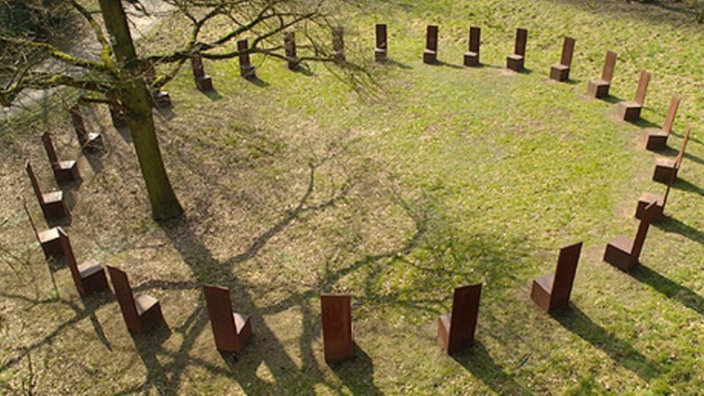 Viele Stühle sind auf einer Wiese in einem großen Kreis aufgestellt und schließen einen Baum ein