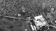 Schwarzweißaufnahme vom 16.08.1969: Die Bühne des Woodstock-Festivals und Tausende Fans, Blick aus der Vogelperspektive