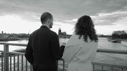 Ein Pärchen steht nach Norden schauend auf der Sevrinsbrücke in Köln, von hinten fotografiert, schwarzweiß.