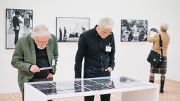 Zwei Personen mit grauen Haaren sind über eine Vitrine gebeugt.