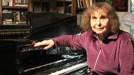 Die Komponistin Sofia Gubaidulina sitzt an ihrem schwarzen Flügel und lächelt. 