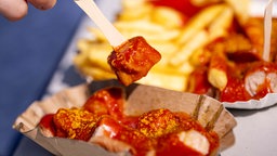 Eine Person spießt ein Stück Currywurst aus einer Schale auf. 