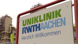 Auf einem Schild steht Uniklinik RWTH Aachen Herzlich Willkommen, dahinter ist ein Gebäude zu sehen.