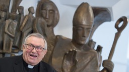 Kardinal Lehmann wird 80: Ein lachender Kardinal am Bildrand, im Hintergrund christliche Bronzestatuen.