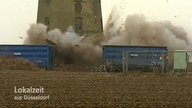 Am unteren Teil des Wasserturms explodiert eine Sprengladung, vor dem Turm stehen Container zum Schutz