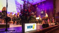 Ein Leuchtschild mit der Aufschrift "WDR 3" vor einem großen, dekorativen Bluemenstrauß, im Hintergrund die Bielefelder Philharmoniker.