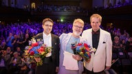 Wolfgang Huber (links) und Alexander Kalajdzic (rechts) erhalten von WDR 3 Moderator Jörg Lengersdorf (Mitte) Blumensträuße.