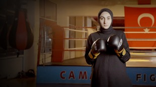 Frau mit Kopftuch steht vor einem Boxring in entsprechender Pose und mit Boxhandschuhen.