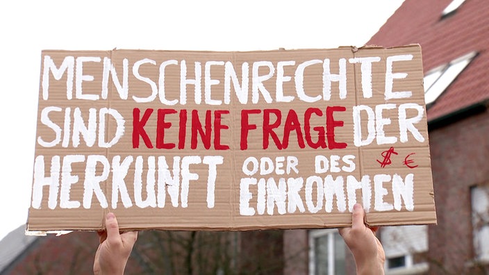 Zwei Hände halten ein Schild mit der Auschrift "Menschenrechte sind keine Frage der Herkunft oder des Einkommens" hoch.