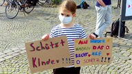 Ein Kind mit Mundschutz bei einer Kundgebung: Das Mädchen hält zwei Plakate hoch mit den Aufschriften "Schützt Kinder" und "Alle Kinder haben das Recht auf Schutz"