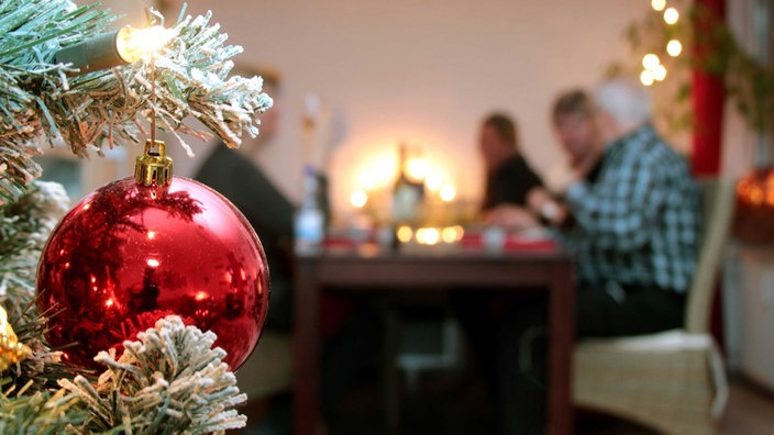 Im Vordergrund hängt eine Christbaumkugel, im Hintergrund sitzt eine Familie beim Weihnachtsessen.