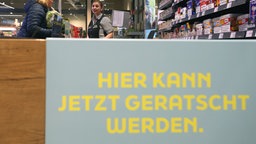 Es ist eine Supermarktkasse zu sehen. Im Vordergrund steht ein Schild mit der Aufschrift: Hier kann jetzt geratscht werden. 