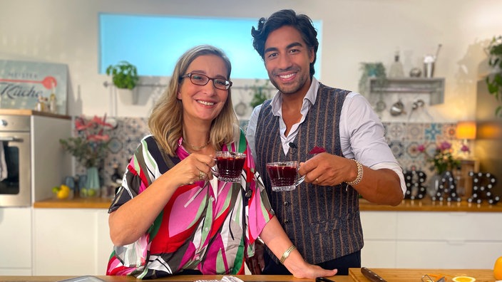 Moderatorin Yvonne Willicks und Bartender Nic Shanker stehen im Kochstudio und halten Drinks hoch.