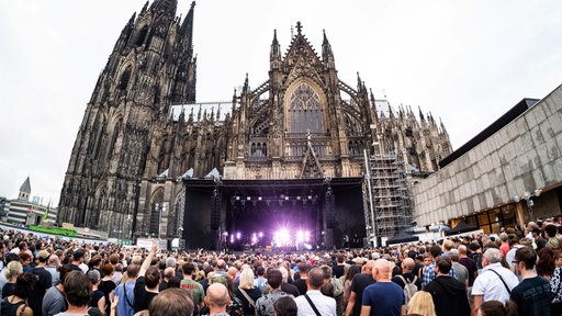 Eine große Menschenmenge steht auf dem Roncalli-Platz zur großen Bühne im Hintergrund gewandt. Hinter der Bühne ragt das Seitenschiff des Kölner Doms in den Himmel.