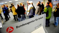 Menschen stehen in einer Reihe, davor ein Schild mit der Aufschrift "Bundesagentur für Arbeit"