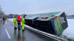 Ein Linienbus liegt auf der Seite im Straßengraben; es liegt Schnee; im Hintergrund sieht man ein Feuerwehrauto; neben dem Bus stehen Männer in Neonkleidung