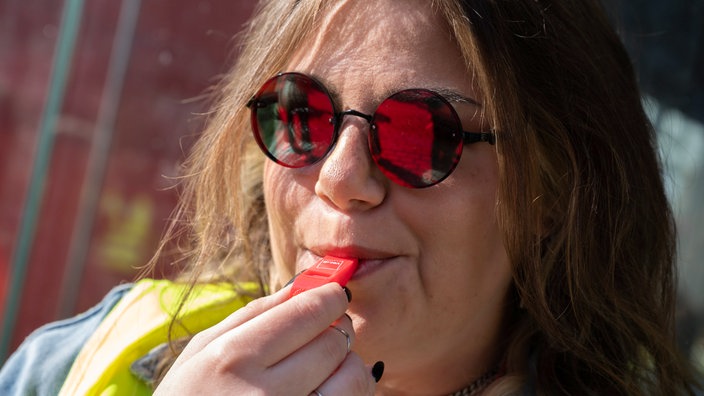 Eine Frau mit Sonnenbrille und Warnweste pustet in eine Pfeife