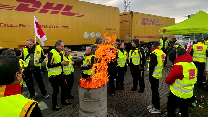 Mitarbeiter und Mitarbeiterinnen der DHL und Verdi streiken und stehen an einem Lagerfeuer. Im Hintergrund sind Lastwägen der DHL zu sehen.